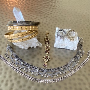 May include: An assortment of gold and silver-toned jewelry. The image features multiple gold bangles, a silver necklace, earrings, and a headpiece. The jewelry is displayed on a light-colored surface, with a crystal and a white rock in the background.