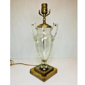 May include: A clear glass table lamp base shaped like a two-handled urn. The lamp features a square brass base with a textured edge, a brass collar, and a brass socket assembly at the top. A thin electrical cord extends from the side of the base.