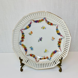 May include: A white porcelain serving plate with a pierced rim and gold trim. The plate is decorated with colorful floral garlands and scattered flower clusters. The plate is displayed on a decorative brass stand.