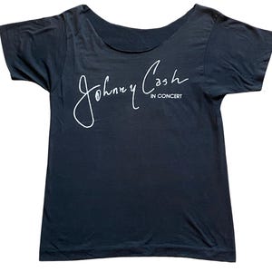 K&ouml;nnte beinhalten: Ein schwarzes T-Shirt mit weitem Ausschnitt. Das Shirt zeigt den wei&szlig;en kursiven Text "Johnny Cash" &uuml;ber den Worten "IN CONCERT". Das Shirt hat kurze &Auml;rmel und eine lockere Passform. Der Stoff wirkt weich und leicht abgenutzt.