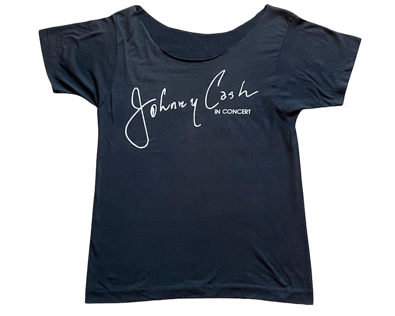 K&ouml;nnte beinhalten: Ein schwarzes T-Shirt mit weitem Ausschnitt. Das Shirt zeigt den wei&szlig;en kursiven Text "Johnny Cash" &uuml;ber den Worten "IN CONCERT". Das Shirt hat kurze &Auml;rmel und eine lockere Passform. Der Stoff wirkt weich und leicht abgenutzt.