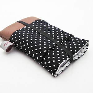 Può includere: Una custodia per telefono in tessuto a pois bianchi e neri, con una patta in tessuto marrone e una chiusura elastica nera. La custodia ha una piccola etichetta con il testo "Arte" su di essa.