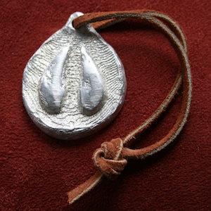 May include: Silver-colored pendant with a textured surface and two raised, teardrop-shaped elements. The pendant is attached to a brown leather cord, knotted at the end. The background is a deep red color.