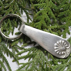 May include: A silver-colored metal keychain with a decorative floral design on the end. The keychain has a large, round ring for attaching keys. The background is a bed of green foliage.