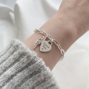 Può includere: Un braccialetto d'argento con un design a catena a clip. Il braccialetto presenta un ciondolo a forma di cuore con inciso "Love You x" e un ciondolo ad ala. Il braccialetto è mostrato su un polso.