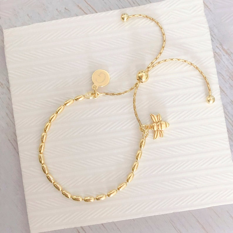 May include: A gold beaded bracelet with a dragonfly charm and a small round charm with a letter 'C' engraved on it. The bracelet has an adjustable sliding clasp.