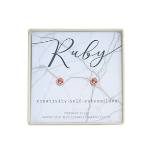 May include: A small white box with a marble pattern. The box contains a pair of sterling silver stud earrings with pink gemstones. The box has the word "Ruby" written in cursive on the top and the words "creativity|self-esteem|love" written on the bottom. The box also has the words "STERLING SILVER" and "www.thelittlekeepsakecompany.co.uk" printed on the bottom.