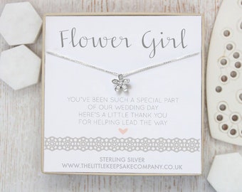 May include: A silver necklace with a small flower charm, packaged in a white box with a gray lace border. The box has the text "Flower Girl" and a message thanking the flower girl for helping lead the way at the wedding. The text "Sterling Silver" and the website address "www.thelittlekeepsakecompany.co.uk" are also printed on the box.