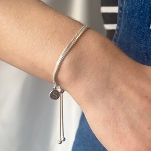 Könnte beinhalten: Silbernes Armband mit Fischgrätmuster. Es hat einen runden Anhänger mit dem Buchstaben "S" und hängenden silbernen Perlen. Das Armband wird am Handgelenk getragen.