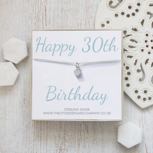 Op de afbeelding: Een witte cadeaudoos met een zilveren ketting en een klein hartvormig bedeltje. De doos heeft de tekst "Happy 30th Birthday" in blauwe letters en de tekst "Sterling Silver www.thelittlekeepsakecompany.co.uk" in zwarte letters.