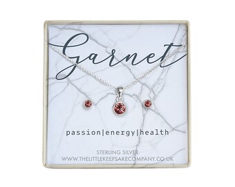 Könnte beinhalten: Ein Collier und Ohrring-Set aus Sterlingsilber mit einem Granat in einer weißen Box mit Marmormuster. Die Box trägt die Aufschriften "Garnet", "passion|energy|health", "Sterling Silver" und "www.thelittlekeepsakecompany.co.uk".