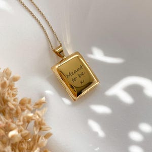 Op de afbeelding: Gouden ketting met een vierkant medaillon. Het medaillon is gegraveerd met de woorden "Meant to be" en een handgeschreven "x".