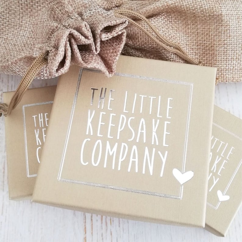 May include: Two small, brown boxes with silver foil accents. The boxes have the text "The Little Keepsake Company" printed on them. The boxes are on a white wooden surface.