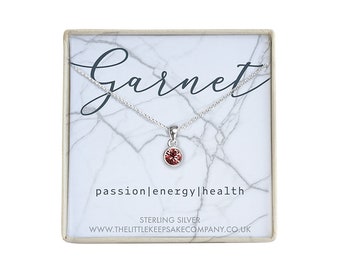 Könnte beinhalten: Eine silberne Halskette mit einem kleinen roten Granat-Anhänger in einer weißen Schachtel mit den Worten "Garnet", "passion|energy|health", "STERLING SILVER" und "www.thelittlekeepsakecompany.co.uk".