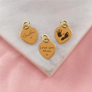 Könnte beinhalten: Drei goldene herzförmige Anhänger mit verschiedenen Gravuren. Ein Anhänger sagt "Oma" mit einem Herzsymbol, ein anderer sagt "Ich liebe dich Mama x", und der dritte Anhänger hat einen Handabdruck und einen Fußabdruck.