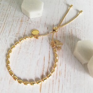 May include: A gold adjustable bracelet with a bee charm and a round disc charm. The bracelet is made of small, oval-shaped beads.