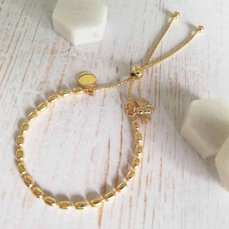 May include: A gold adjustable bracelet with a bee charm and a round disc charm. The bracelet is made of small, oval-shaped beads.