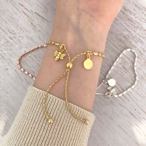 May include: Two gold chain bracelets with adjustable sliding knots. One bracelet has a bee charm and a round disc charm. The other bracelet has a round disc charm.