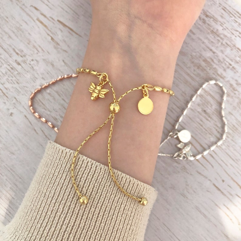 May include: Two gold chain bracelets with adjustable sliding knots. One bracelet has a bee charm and a round disc charm. The other bracelet has a round disc charm.