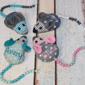 Könnte beinhalten: Vier graue und blaue Stoffmäuse mit bestickten Namen Avery, Eliana und zwei weiteren. Jede Maus hat einen Schwanz aus einer Schnur mit bunten Perlen.