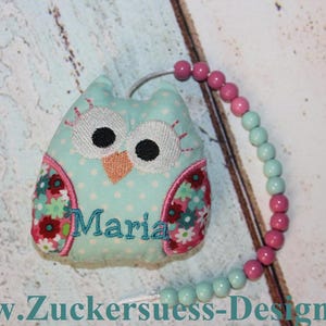 Könnte beinhalten: Ein hellblaues Plüschtier in Form einer Eule mit rosa und grünen Blumenapplikationen. Die Eule hat gestickte Augen und einen Schnabel. Das Spielzeug ist an einer Schnur aus rosa und hellblauen Perlen befestigt. Der Name "Maria" ist auf die Brust der Eule gestickt.