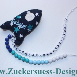 May include: A blue and white rocket-shaped fabric toy with the name "Gabriel" embroidered on it. A string of alphabet beads in blue, turquoise, and white is attached to the toy.
