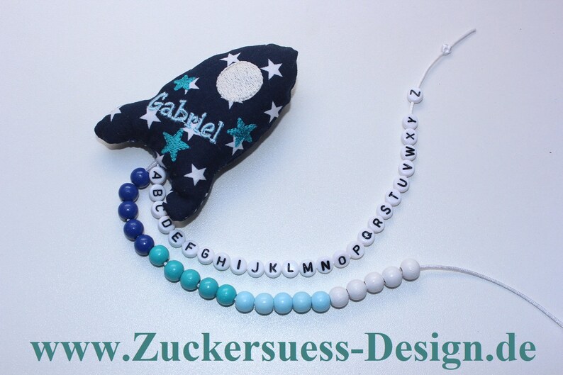 May include: A blue and white rocket-shaped fabric toy with the name "Gabriel" embroidered on it. A string of alphabet beads in blue, turquoise, and white is attached to the toy.