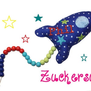 May include: A blue rocket-shaped plush toy with white polka dots and stars. The toy has a name "Phil" embroidered on it and is attached to a colorful beaded string. The text "Zuckers&uuml;&szlig;" is written below the toy.