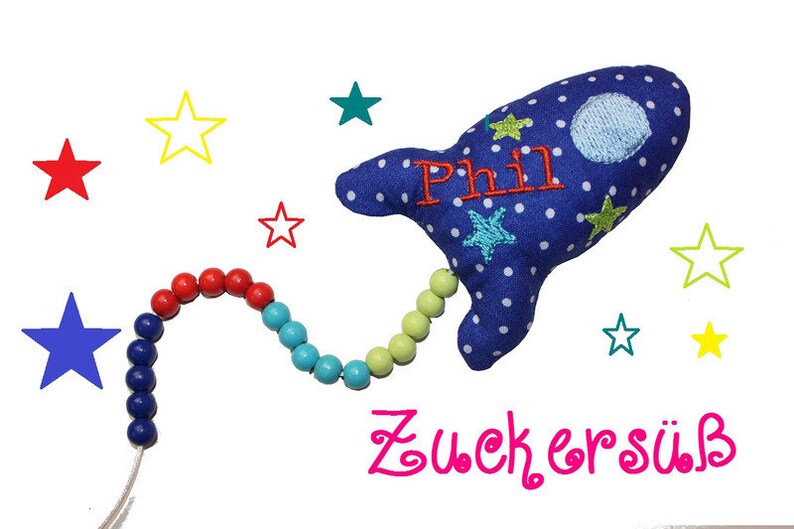 May include: A blue rocket-shaped plush toy with white polka dots and stars. The toy has a name "Phil" embroidered on it and is attached to a colorful beaded string. The text "Zuckers&uuml;&szlig;" is written below the toy.