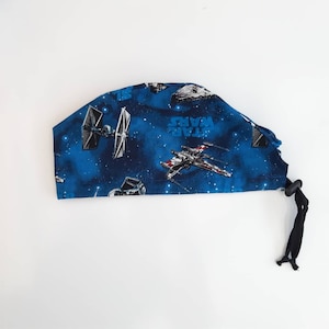 Könnte beinhalten: Blaue OP-Mütze mit Star Wars-Motiv. Die Mütze zeigt ein sich wiederholendes Muster aus X-Wings, TIE-Jägern und dem Star Wars-Logo auf dunkelblauem Hintergrund mit weißen Sternen.