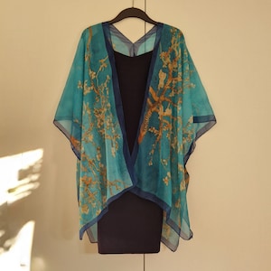 May include: A teal blue kimono with a floral print of a tree with yellow flowers. The kimono is hanging on a hanger against a white wall.