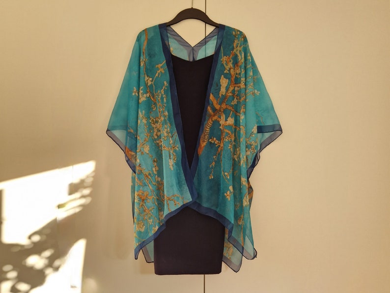 May include: A teal blue kimono with a floral print of a tree with yellow flowers. The kimono is hanging on a hanger against a white wall.