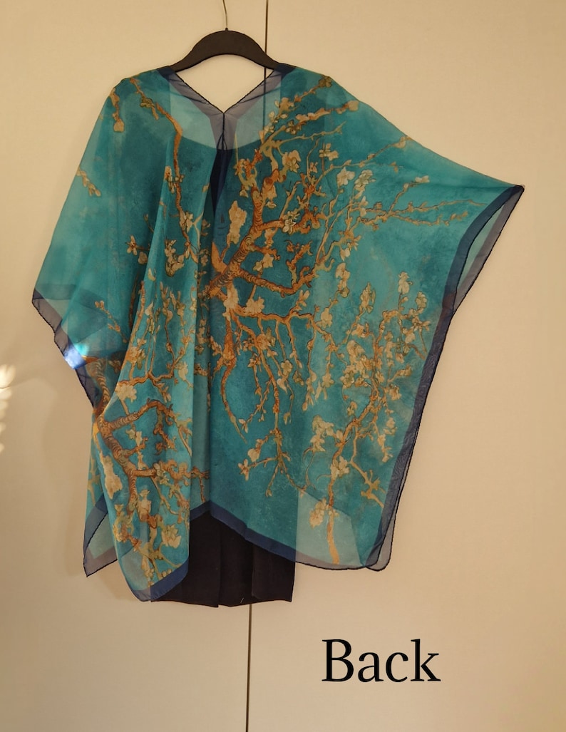 May include: A turquoise blue sheer shawl with a printed design of a tree with white blossoms. The shawl is draped over a hanger and has a V-neckline. The back of the shawl is shown in the image. The text "Back" is printed below the shawl.