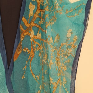 May include: A turquoise blue sheer scarf with a printed design of a tree with white blossoms. The scarf has a dark blue trim.