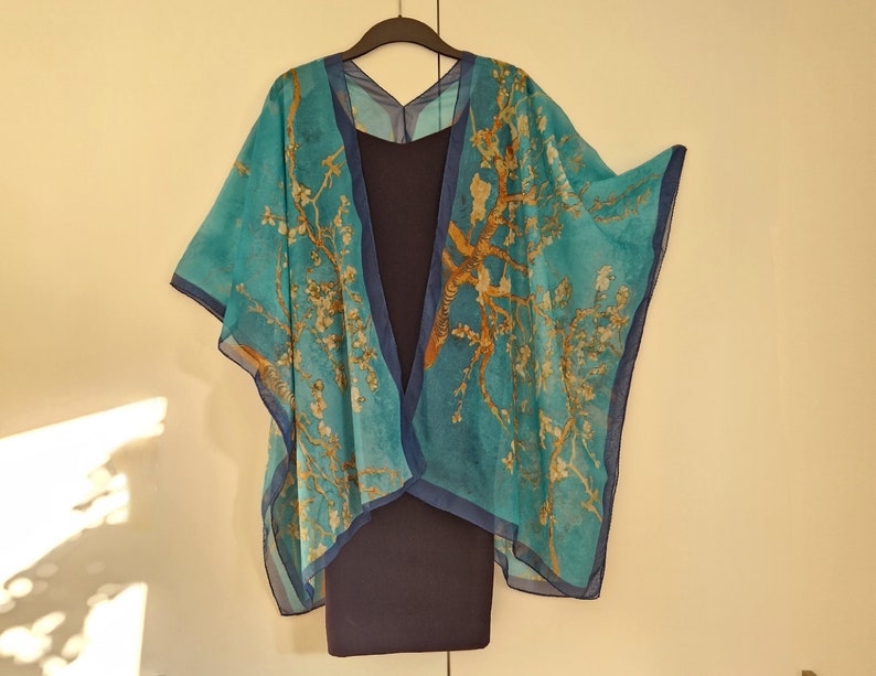 May include: A turquoise blue sheer kimono with a floral and bird print. The kimono has a V-neck and is open in the front. It is hanging on a black hanger.