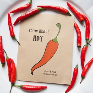 以下が含まれることがあります： 白い皿の上に赤い唐辛子が置かれ、その上に「some like it HOT」と書いてある茶色の紙のカードが置かれている。カードの下には「Jenny & Mark 17th September 2014」と書かれている。カードには赤い唐辛子の漫画が描かれている。