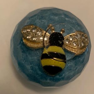Könnte beinhalten: Blaues, facettiertes Dekorationsobjekt mit einer goldfarbenen Biene. Die Biene hat schwarz-gelbe Streifen und mit Kristallen besetzte Flügel. Dieser Artikel ist wahrscheinlich ein dekoratives Stück, möglicherweise ein Briefbeschwerer oder Ornament.