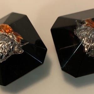 May include: Pair of black, faceted square earrings. Each earring features a silver-colored sculpted design with an orange bow accent. The earrings are displayed on a light-colored surface.