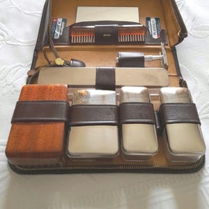 Peut inclure: Une trousse de toilette de voyage vintage en cuir marron, ouverte pour afficher son contenu. Le kit comprend un peigne, un miroir, un rasoir et plusieurs contenants. L'intérieur est doublé d'un matériau marron clair et la trousse se ferme avec une fermeture éclair.