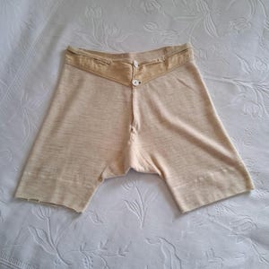 May include: Cream-coloured vintage underwear shorts with a button-up front and a contrasting tan waistband. The shorts have a ribbed hem and a classic design. The fabric appears to be a soft, knit material.