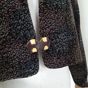 Può includere: Giacca nera con un motivo a foglie colorate. La giacca ha due grandi bottoni rotondi marroni con un centro giallo. Le maniche sono lunghe e il tessuto ha una consistenza morbida e vellutata. Un capo di abbigliamento esterno elegante.