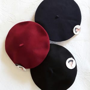 May include: Three beret hats in burgundy, navy blue, and black. Each hat is round with a small, centered nub. A small tag with a portrait is attached to each beret. The hats are displayed on a white, textured surface.
