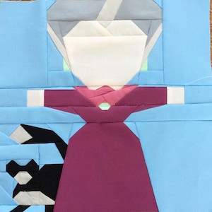 May include: A paper pieced quilt block featuring a woman in a red dress with a white face and gray hair. The background is light blue.
