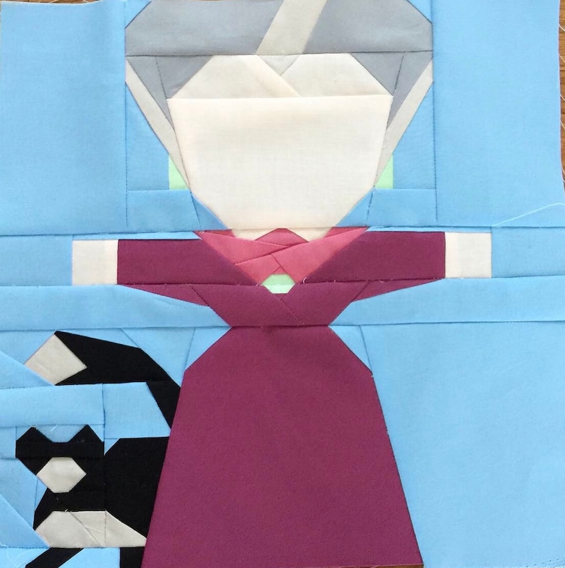May include: A paper pieced quilt block featuring a woman in a red dress with a white face and gray hair. The background is light blue.
