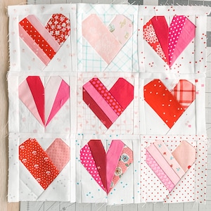 May include: Nine patchwork hearts arranged in a 3x3 grid. Each heart is made from different red and pink patterned fabrics. The hearts are sewn onto a white background.