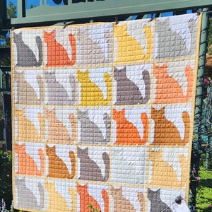 Op de afbeelding: Een quilt met een patchwork-ontwerp met een herhalend patroon van katten in verschillende kleuren, waaronder oranje, geel, bruin en grijs. De quilt is gemaakt van vierkanten met een witte achtergrond en een gestikte textuur.