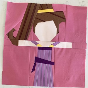 May include: A pink fabric quilt block with a purple and yellow dress and brown hair. The quilt block is made using the paper piecing technique.