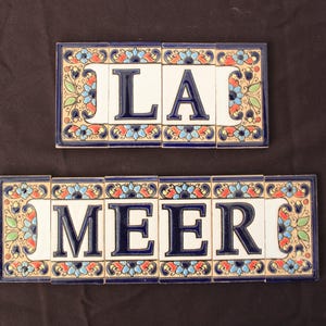 Op de afbeelding: Twee rechthoekige keramische tegels met de letters "LA" en "MEER" in donkerblauw. De tegels zijn wit met een blauwe rand en bloemmotieven in oranje, blauw en groen.