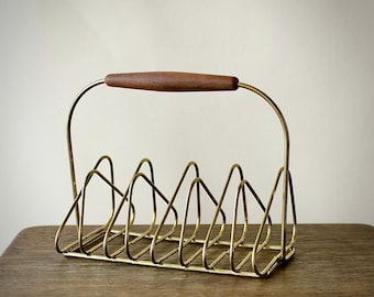 May include: A vintage-style magazine rack with a gold-toned metal frame and a wooden handle. The rack features a series of curved dividers to hold magazines or files. The handle is a dark brown color.