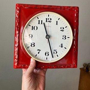 May include: A square wall clock with a glossy red ceramic frame featuring a textured, uneven border. The clock face is off-white with black Arabic numerals, a gold-toned circular bezel, and black hands. The text ZentRa is printed in black.